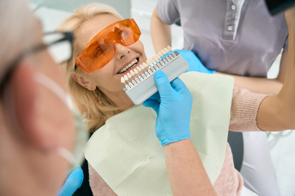 A female patient lies in the treatment chair with orange protective glasses on. Her dentist is comparing her tooth size, shape, and color/tone with a set of implants. They just finished talking about the teeth implant's price in Overland Park.