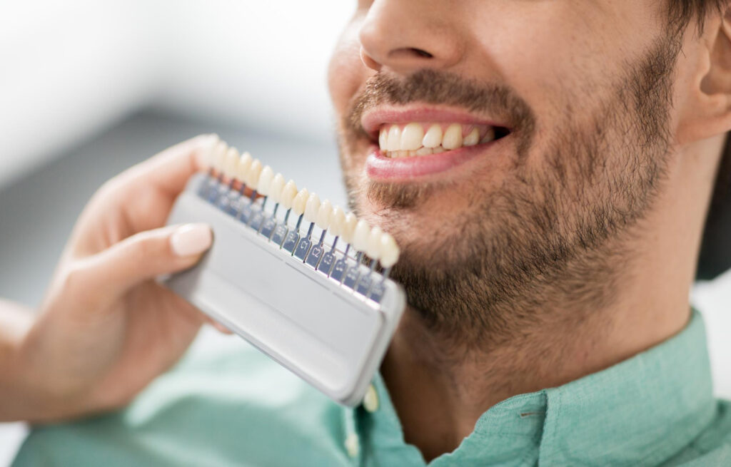 Patient smiling while a dentist holds up a selection of crowns to his teeth. This is part of a consultation and overall discussion regarding dental implant cost in Overland Park.