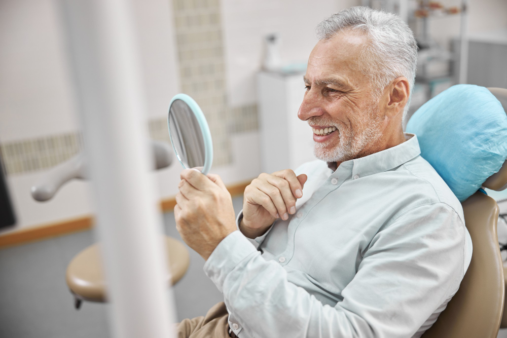 An older man smiles at his reflection in a handheld mirror at our office after getting professional teeth whitening near Lenexa.